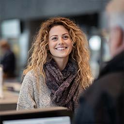 A woman with curly hair and a scarf smiles while speaking to someone across a counter in an indoor setting.