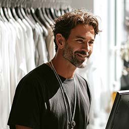 A man with curly hair and a beard, wearing a black t-shirt and necklace, stands in front of a rack of clothes, smiling at a computer screen.